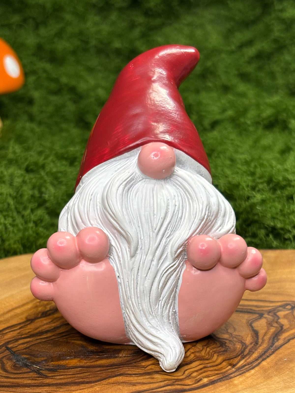A gnome figurine with a red hat, white beard, and pink feet, displayed on a wooden board with green moss and small decorative mushrooms in the background.