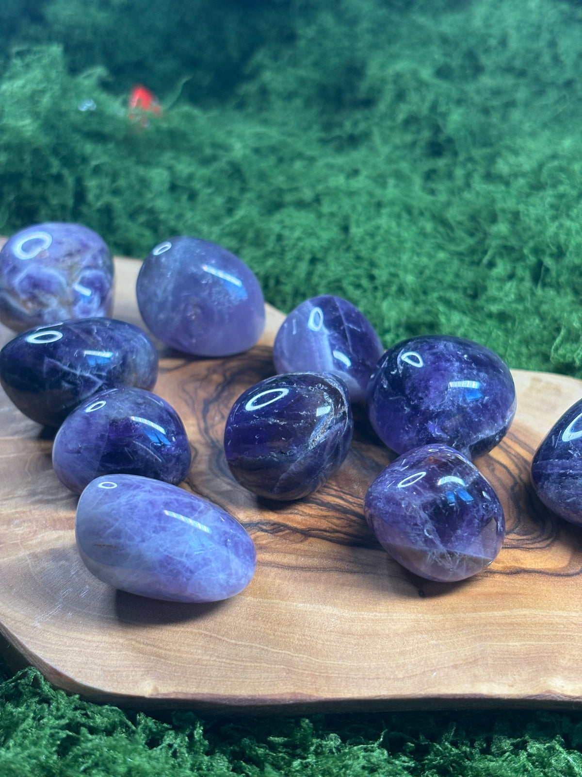 Assorted natural amethyst tumbled stones with unique purple coloring and patterns displayed on wood and green moss.