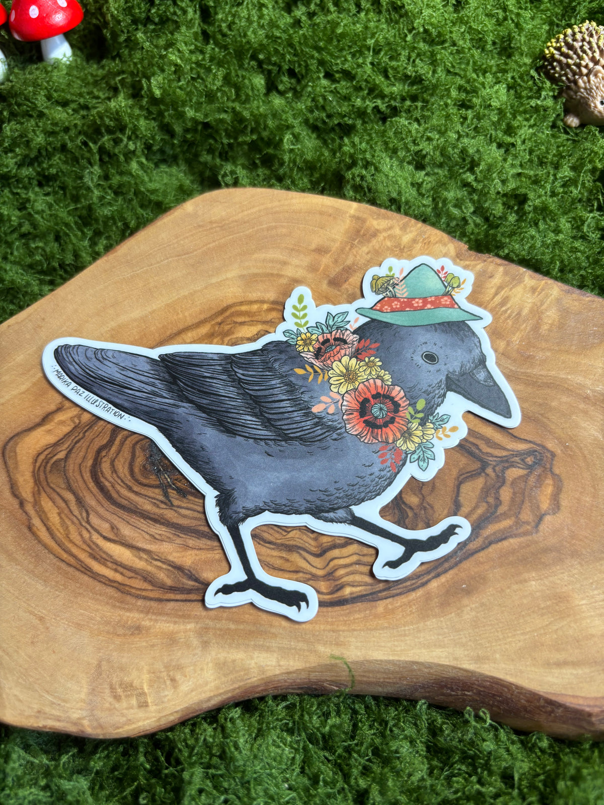 Whimsical corvid sticker with green hat and flowers displayed on wood