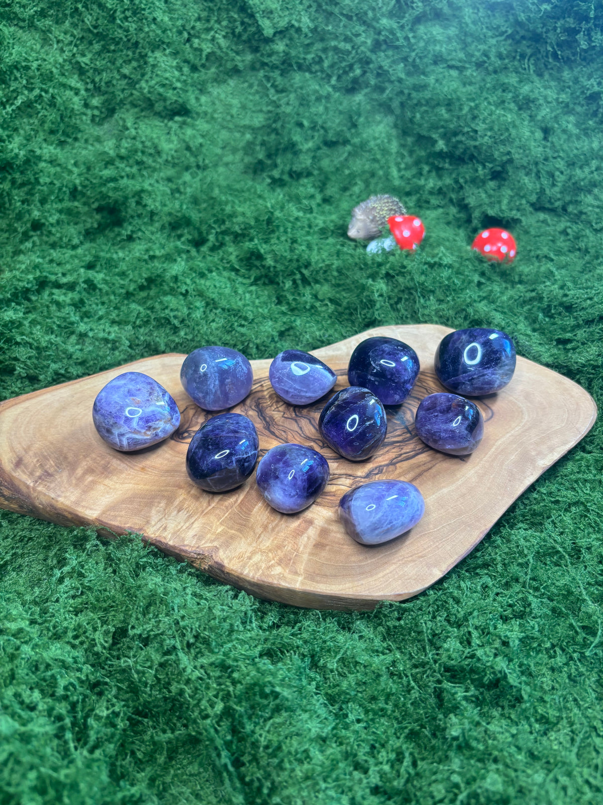 A group of polished purple gemstones arranged on a natural wood slice, set against a green moss background with small decorative mushrooms in the distance.